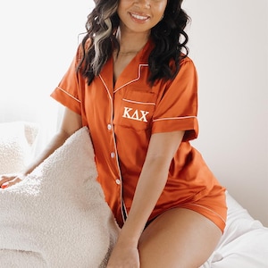 Puede incluir: Una mujer con un pijama de satén color óxido con ribetes blancos y las letras "ΚΔΧ" bordadas en el pecho.