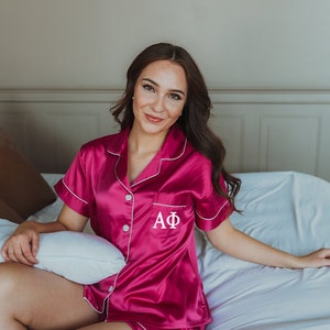 May include: A woman wearing a pink satin pajama set with white piping and the letters "AΦ" embroidered on the shirt. She is sitting on a white bed with a white pillow.