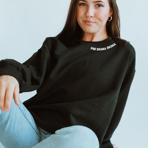 May include: A black long-sleeved sweatshirt with the text "PHI SIGMA SIGMA" embroidered on the neckline. The sweatshirt is being worn by a person sitting with their legs crossed, wearing blue jeans.