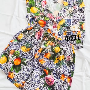 Peut inclure: Un ensemble pyjama blanc et bleu avec un imprimé floral et fruitier. Le haut a un col et des manches courtes. Le short a une taille élastique. L'ensemble présente un motif de lettres grecques brodées en noir qui dit "Phi Sigma II".