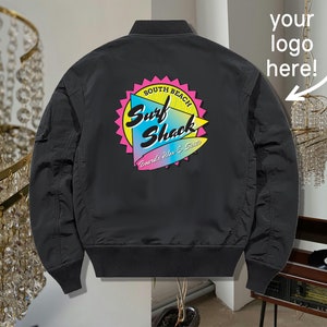 May include: Black bomber jacket with a colorful "Surf Shack" logo on the back. The logo features a surfboard and the text "South Beach" and "Boards, Vibes & Sun".
