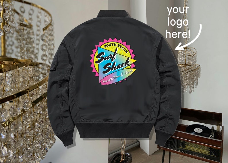 May include: Black bomber jacket with a colorful "Surf Shack" logo on the back. The logo features a surfboard and the text "South Beach" and "Boards, Vibes & Sun".
