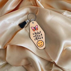 May include: A light brown motel key chain with a black tassel, a pink butterfly, and the text "alpha sigma alpha" in black script. A yellow smiley face is at the bottom of the keychain.