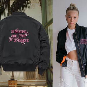 May include: Black bomber jacket with a pink embroidered design on the back that says "Creating the life I deserve".