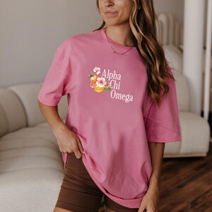 Puede incluir: Una camiseta rosa con el texto "Alpha Chi Omega" y un diseño floral. La camiseta se combina con pantalones cortos marrones. La imagen está tomada en interiores.