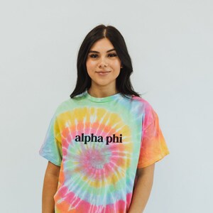 May include: A tie-dye t-shirt with a rainbow swirl pattern and the text "alpha phi" in black lettering.