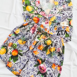 May include: A white and blue patterned pajama set with a short-sleeved top and shorts. The top has a button-up closure and a collar. The shorts have an elastic waistband. The fabric features a floral and fruit design with lemons, peaches, and kiwi. The top has the text "Splkes and Seams" on the front. The shorts have the text "ZITA" on the back.