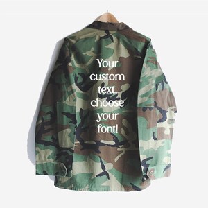 Puede incluir: Una chaqueta verde de camuflaje con el texto "Your custom text choose your font!" impreso en la espalda.