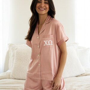 May include: A pink satin pyjama set with short sleeves and shorts. The top has a white button fastening and a white pocket with the letters "XO" embroidered on it.