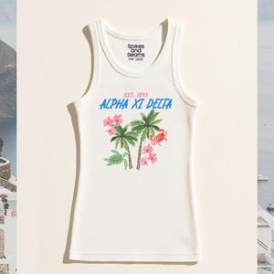 Op de afbeelding: Witte geribbelde tanktop met een tropisch ontwerp. Het heeft palmbomen, roze bloemen en een krab. De tekst "ALPHA XI DELTA" is in blauw, met "EST. 1893" in roze. De top heeft het "Spikes and Seams" logo.
