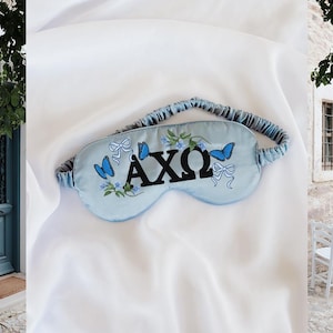 May include: A light blue sleep mask with the Greek letters AXΩ embroidered in black. The mask features blue butterflies, bows, and floral accents. The elastic band is blue and white striped.