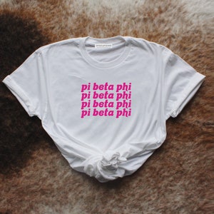 Puede incluir: Camiseta blanca con el texto "pi beta phi" en rosa, repetido cuatro veces. La camiseta está anudada en la parte inferior. La marca "SPIRIT OF JEANS" está impresa en la parte superior.