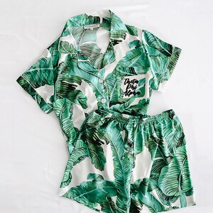 Puede incluir: Conjunto de pijama blanco con estampado de hojas tropicales verdes. La parte superior tiene cuello abotonado y mangas cortas. Los pantalones cortos tienen cintura elástica. El bolsillo de la parte superior tiene el texto "Theta Phi Alpha" bordado.