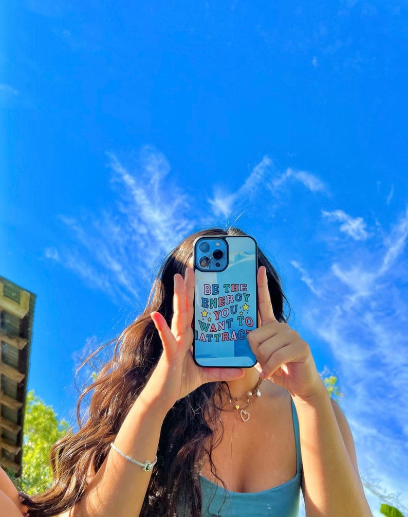 May include: A person is holding a blue and white phone case with the text "Be the energy you want to attract" in colourful letters. The phone case has a white background with a blue and white design.