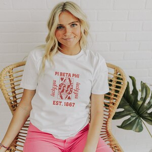 Puede incluir: Camiseta blanca con un gráfico rojo de una carta de reina de corazones con el texto "Pi Beta Phi" y "lucky you" a cada lado de la carta. El texto "EST. 1867" está debajo de la carta.