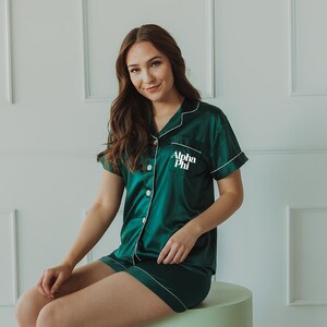 以下が含まれることがあります： 胸に「Alpha Phi」と刺繍された、白いパイピングが付いた緑色のサテンパジャマを着た女性です。