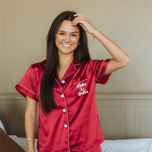 May include: A woman wearing a red satin pajama set with white piping. The top has a pocket with the text "Alpha Xi Delta" embroidered on it.