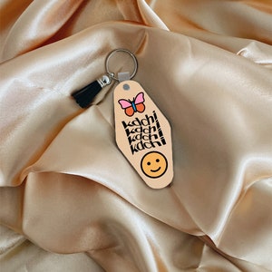 Puede incluir: Un llavero de motel beige con una mariposa rosa, texto negro que dice "kachi" cuatro veces y una carita sonriente amarilla.