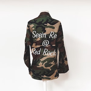 May include: A camouflage green, brown, and beige utility jacket with the text "Seein Red @ Red Rocks" printed on the back.