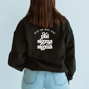 May include: A black sweatshirt with the text "Wish you were here" and "Phi Sigma Sigma" in white lettering on the back. The sweatshirt is being worn by a person with long brown hair and blue jeans.