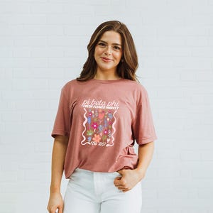 Puede incluir: Camiseta de color rosa polvoriento con un diseño floral y el texto "pi beta phi FRESH FLOWER MARKET Est. 1867". La camiseta es de material suave y tiene mangas cortas.
