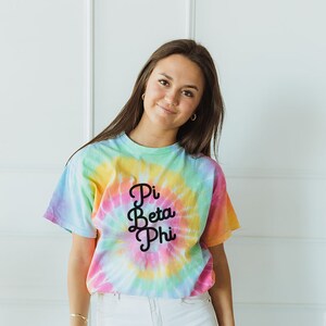 Puede incluir: Una mujer con una camiseta de tie-dye con el texto "Pi Beta Phi" en letras negras. Lleva pantalones vaqueros blancos y tiene el pelo largo y castaño.