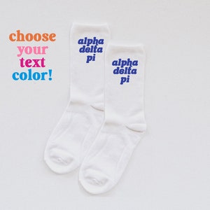 May include: Two white socks with the text "alpha delta pi" in blue lettering. The text "choose your text color!" is written in orange, pink, and blue.