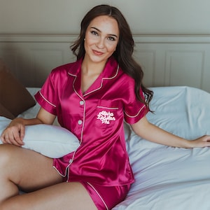 Puede incluir: Una mujer lleva un pijama de satén rosa con ribetes blancos y el texto "Alpha Epsilon Phi" bordado en el pecho.