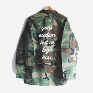 Puede incluir: Una chaqueta utilitaria de color verde camuflaje con un mensaje bordado en blanco en la parte posterior que dice "your custom text or logo here".