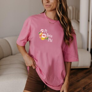 Peut inclure: Un t-shirt rose avec le texte "Pi Beta Phi" et un motif floral. Le t-shirt est associé à un short marron. L'image montre une femme portant le t-shirt et le short.
