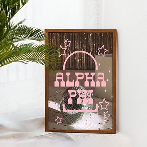 May include: A framed mirror with a pink "ALPHA PHI" design and a disco ball in the reflection. The mirror is surrounded by a wooden frame and has a background of gold glitter and stars.