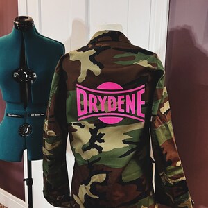 Puede incluir: Chaqueta de camuflaje con un logotipo rosa que dice "Drydene" en la espalda.