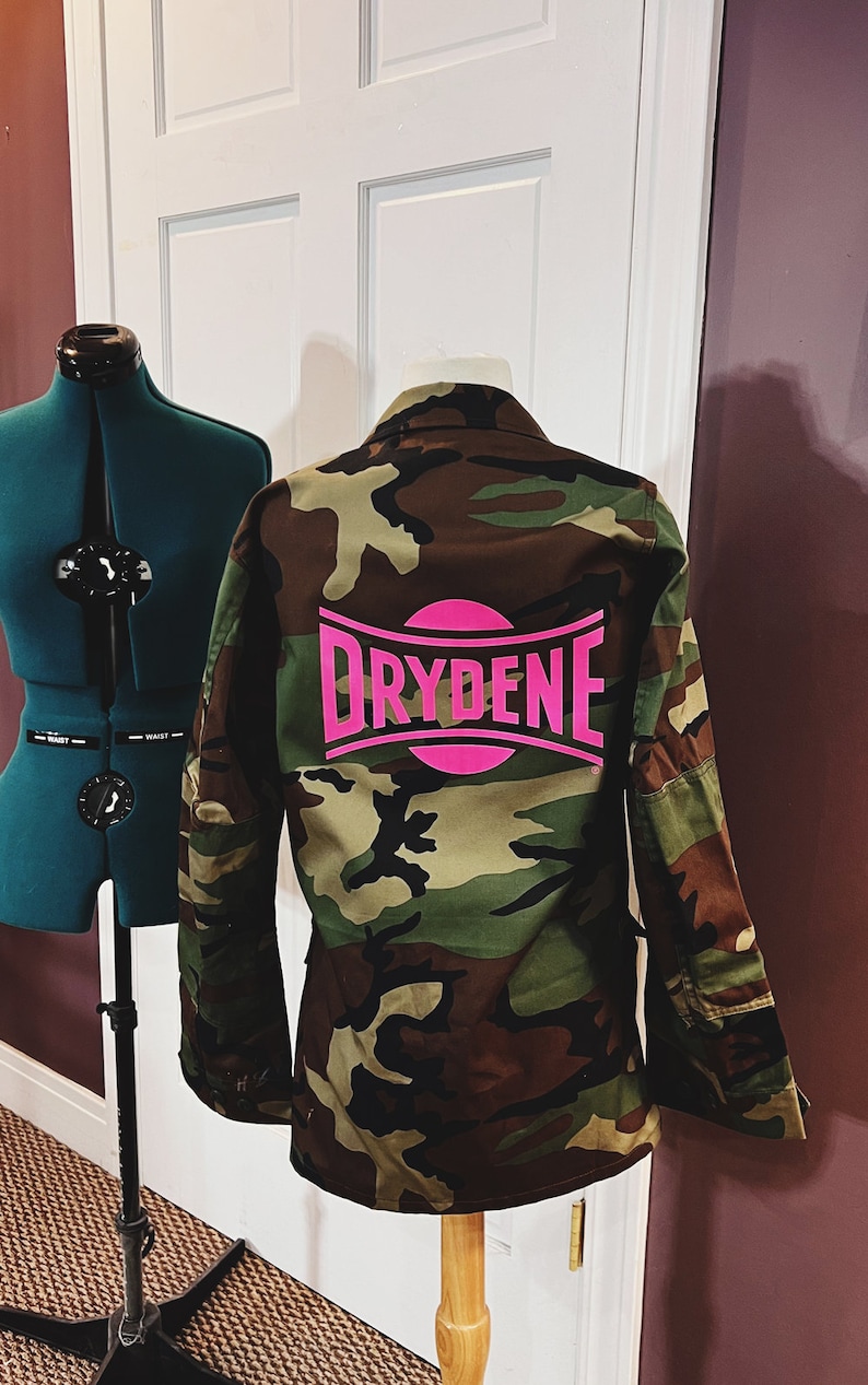 Puede incluir: Chaqueta de camuflaje con un logotipo rosa que dice "Drydene" en la espalda.