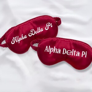 Puede incluir: Dos antifaces de satén rojo con texto bordado en blanco, "Alpha Delta Pi", en cada antifaz.