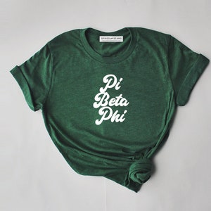 Puede incluir: Camiseta verde con texto blanco que dice "Pi Beta Phi" en una fuente retro.