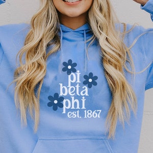 Könnte beinhalten: Hellblauer Kapuzenpullover mit einem weißen Grafikdesign mit dem Text "pi beta phi est. 1867" umgeben von blauen Blumen.