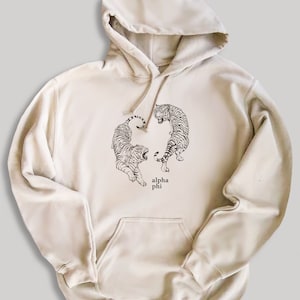 May include: A beige hoodie with a black and white illustration of two tigers facing each other. The text "alpha phi" is printed below the tigers.