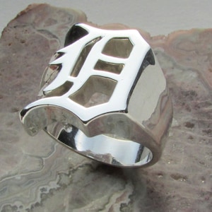 Puede incluir: Un anillo de plata pulida con un diseño audaz y estilizado de la letra "D". El anillo tiene una banda ancha y una superficie lisa y reflectante, que muestra una estética moderna y geométrica. El anillo es una pieza llamativa.