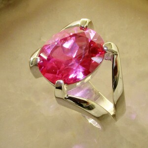 May include: A silver ring featuring a large, oval-cut, bright pink gemstone. The gemstone is set in a modern, minimalist design with a polished silver band. The ring is displayed on a light-colored surface.