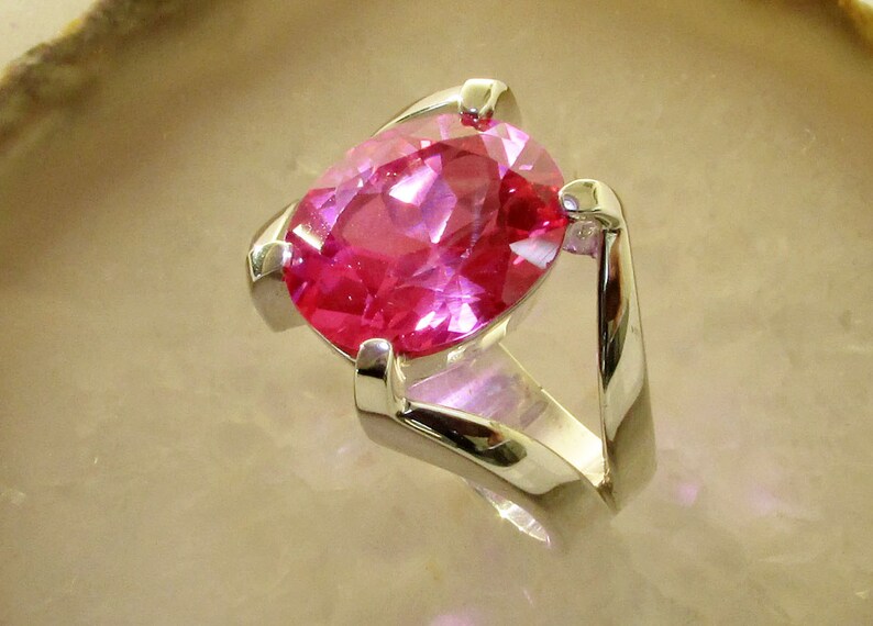 May include: A silver ring featuring a large, oval-cut, bright pink gemstone. The gemstone is set in a modern, minimalist design with a polished silver band. The ring is displayed on a light-colored surface.