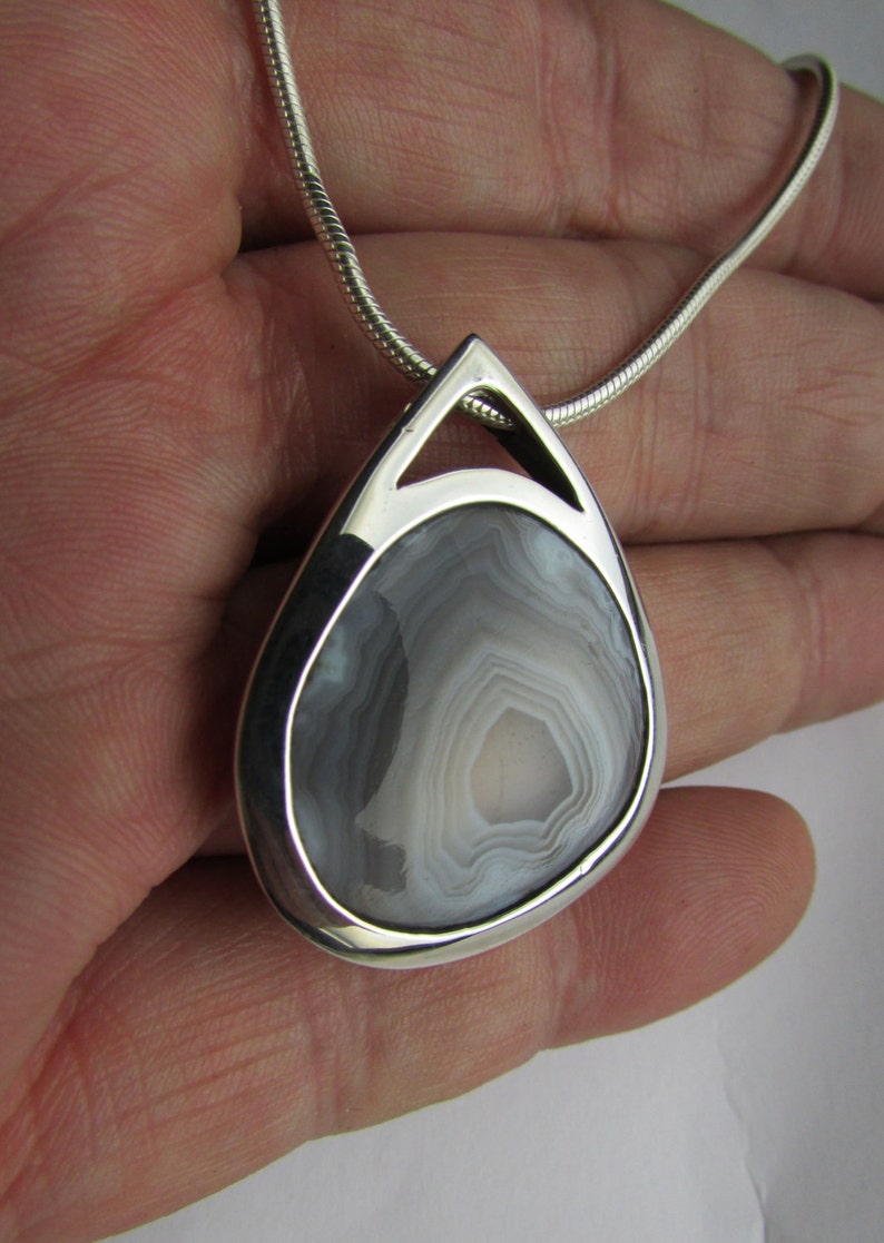 May include: A teardrop-shaped pendant with a polished silver frame, showcasing a gray and white agate stone. The stone has concentric bands of color. The pendant hangs from a silver snake chain. Jewelry.
