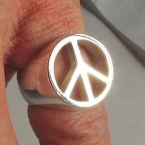 May include: A silver ring featuring a peace symbol design. The ring has a polished, reflective surface and a circular face with the iconic peace sign cut out. The ring is a symbol of peace and unity.