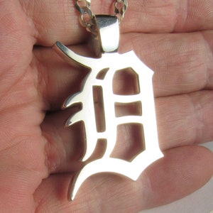 May include: A silver pendant in the shape of the letter "D" in a gothic font, hanging from a silver chain. The pendant is highly polished and reflects light. The chain is a curb style. The pendant is approximately 2 inches in height.