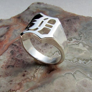 May include: A silver ring with a cut-out design. The ring has a rectangular face with a stylized letter "D" cut out. The ring is set on a multi-colored, patterned surface.