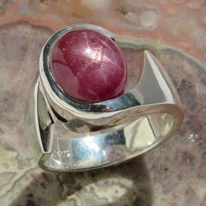 May include: A sterling silver ring featuring a large, oval-shaped, deep red gemstone. The ring has a modern, sculptural design with a polished finish. The gemstone is the focal point, set in a bezel setting.