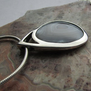 May include: A teardrop-shaped pendant with a polished silver frame. The pendant features a gray and white banded stone, suspended from a silver chain. The stone's pattern is visible, and the pendant is set against a neutral background.