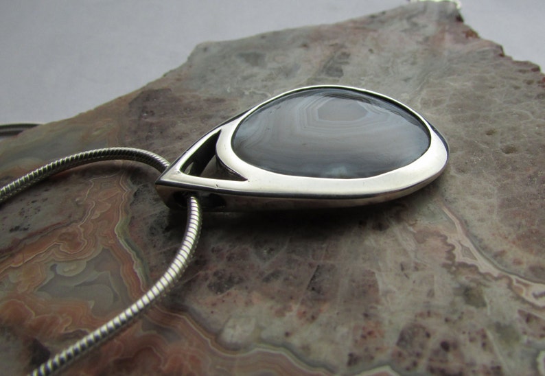 May include: A teardrop-shaped pendant with a polished silver frame. The pendant features a gray and white banded stone, suspended from a silver chain. The stone's pattern is visible, and the pendant is set against a neutral background.