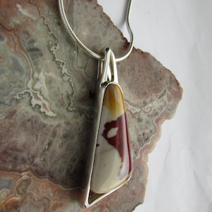 May include: A silver pendant necklace featuring a polished, triangular stone with a mix of colors: cream, burgundy, and yellow. The stone is set in a silver bezel and hangs from a delicate silver chain. The necklace is displayed on a stone surface.