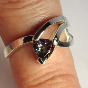 May include: A silver ring with a pear-shaped, multi-colored gemstone. The ring has a unique design with a split band and a decorative element that curves around the gemstone. The gemstone displays a range of colors, including green, purple, and blue.