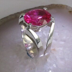 May include: A silver ring featuring a large, faceted, pear-shaped, hot pink gemstone. The ring's band has a unique, open design with curved, flowing lines that cradle the stone. The ring is set against a light background.